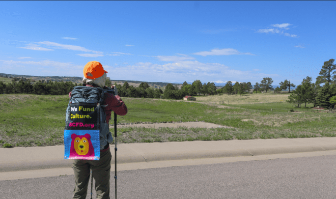 Local naturalist and writer India Wood snaps a photo of a green landscape, an SCFD banner hanging on her backpack.