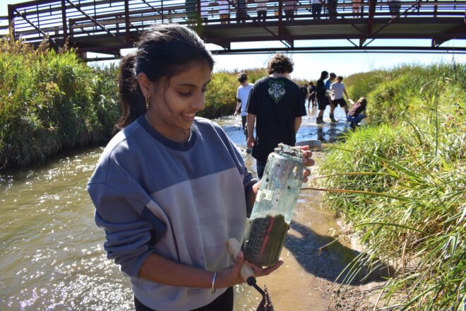 A young girl smiles down at a water bottle filled with river sediment as students wade through a river behind her.